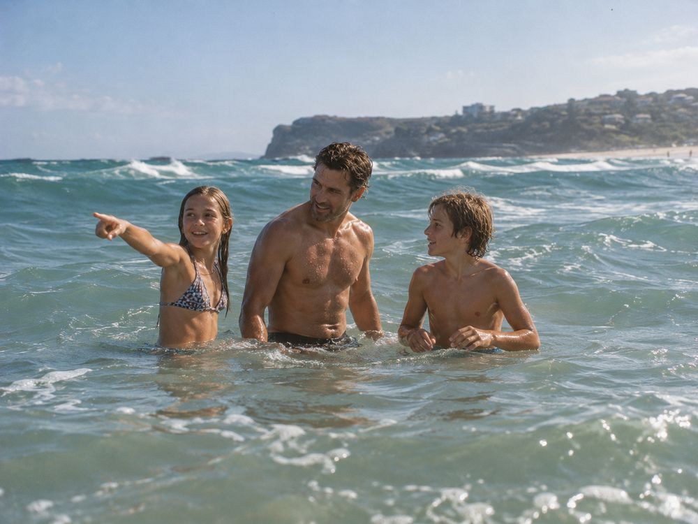 A man in his forties standing waist-deep in the surf with two kids, all talking and laughing
